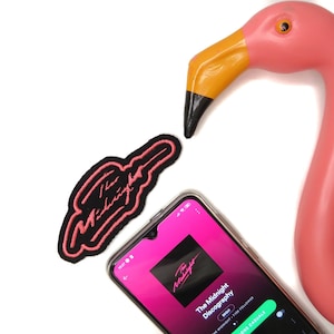 Op de afbeelding: Een roze flamingo-figuur met een zwart en roze geborduurde patch van de band "The Midnight" op een witte achtergrond. Een smartphone die de discografie van de band op Spotify weergeeft is ook zichtbaar.
