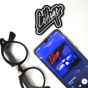 Puede incluir: Un teléfono inteligente que muestra la portada del álbum "Secret Diary" de la banda "College". La portada del álbum presenta un diseño azul y negro con un par de gafas de sol.