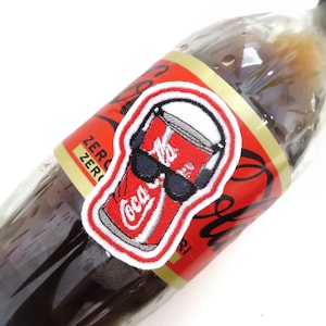 Può includere: Una lattina di Coca-Cola rossa e bianca con occhiali da sole e cuffie, ricamata su una toppa bianca con bordo rosso. La toppa è attaccata a una bottiglia di vetro marrone di Coca-Cola.