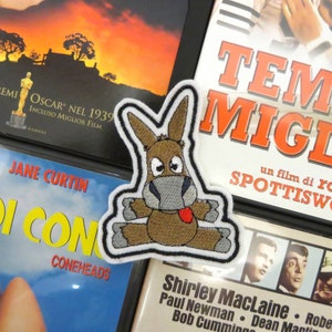 Pode incluir: Um burro castanho com o nariz cinzento e a língua vermelha é bordado em um remendo branco. O remendo é cercado por uma borda branca. O remendo está sentado em cima de uma caixa de DVD com o texto "TEM MIGL" em letras vermelhas.