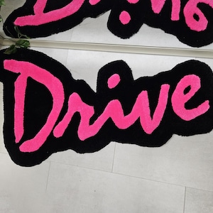 Puede incluir: Una alfombra de bienvenida negra y rosa con la palabra "Drive" escrita en letras grandes de color rosa.