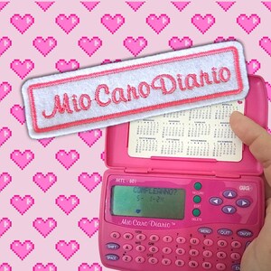 以下が含まれることがあります： 「Mio Caro Diario」と書かれたピンク色の電子日記と、お揃いのパッチ。日記の画面には「COMPLEANNO?」と日付が表示されています。背景には、ピンク色のピクセル化されたハートのパターンがあります。