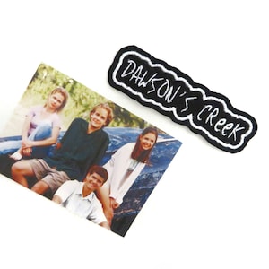 Può includere: Una toppa ricamata in bianco e nero con il testo "Dawson's Creek" in un font bianco corsivo. Sullo sfondo c'è una fotografia di quattro persone.