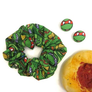 May include: Green and red scrunchie with a Teenage Mutant Ninja Turtles print. Two small green and red buttons with a Teenage Mutant Ninja Turtles face on them. A round, flat, yellow and red pizza.