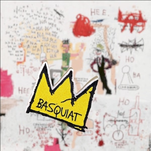 May include: A yellow embroidered crown with black outlines and the word "BASQUIAT" written in black on the front. The crown is on a white background with black and red scribbles.
