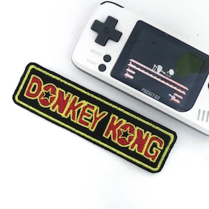 Puede incluir: Un parche bordado negro y amarillo con las palabras "DONKEY KONG" en letras rojas con una estrella amarilla a cada lado del texto. El parche está al lado de una consola de videojuegos portátil blanca con una pantalla negra que muestra el juego Donkey Kong.