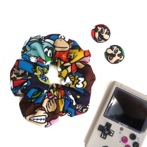 Könnte beinhalten: Ein farbenfroher Haargummi mit einem Muster aus verschiedenen Nintendo-Charakteren, darunter Mario, Luigi, Donkey Kong und Yoshi. Zwei kleine Knöpfe mit Marios Gesicht sind ebenfalls enthalten. Im Hintergrund befindet sich eine graue Game Boy-Handheld-Konsole.