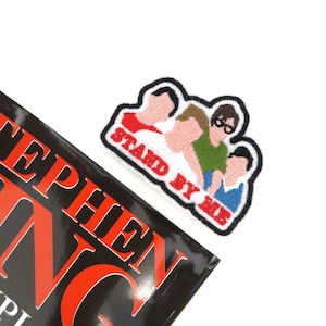 May include: Embroidered patch featuring four cartoon figures with the text "Stand By Me" in red and white. The figures are wearing different coloured shirts and are standing close together.