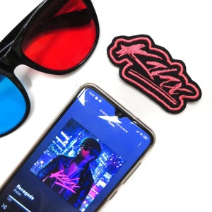 Può includere: Uno smartphone che mostra un'app di lettore musicale con la canzone "Renegade" di Kalax in riproduzione. Lo schermo è blu e il titolo della canzone e l'artista sono bianchi. Il telefono è su una superficie bianca con un paio di occhiali 3D rossi e blu e una toppa ricamata rosa con la parola "Kalax" su di essa.