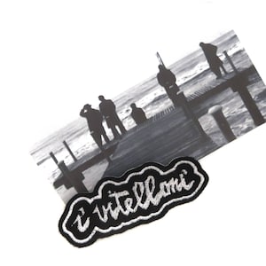 Puede incluir: Foto en blanco y negro de cuatro personas de pie en un muelle de madera sobre el agua. La foto está unida a un parche bordado en blanco y negro que dice "i vitelloni".