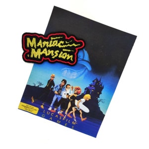 Puede incluir: Un póster de videojuego vintage de Maniac Mansion con un parche bordado en rojo y dorado con el título del juego. El póster presenta una ilustración de dibujos animados de una mansión espeluznante y un grupo de personajes.