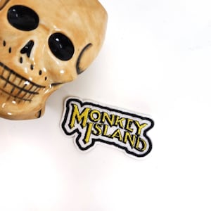 Peut inclure: Un écusson brodé blanc avec un contour noir et des lettres jaunes qui lisent "MONKEY ISLAND".