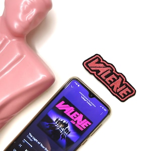 Puede incluir: Un busto de cerámica rosa se encuentra junto a un teléfono inteligente que reproduce música de la banda "Valene and Friends". La canción que se está reproduciendo es "The Light of Your Dreams". Un parche bordado rojo y negro con la palabra "Valene" se encuentra en la esquina superior derecha de la imagen.