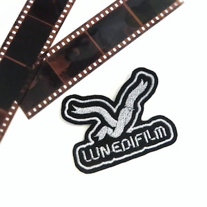 Può includere: Una toppa ricamata in bianco e nero con un design di uccello stilizzato e il testo "LUNEDIFILM" in bianco.