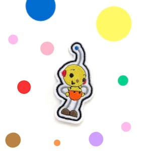May include: Embroidered patch of a yellow cartoon character with red details, an orange nappy, and grey limbs. The patch has a white border with a black outline. The background features colourful polka dots of various sizes and colours.