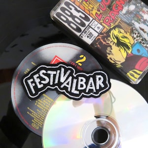 Op de afbeelding: Een geborduurde zwarte en witte patch met de tekst "FESTIVALBAR" in witte letters op een zwarte achtergrond. De patch ligt op een zilveren cd met een zwart midden.