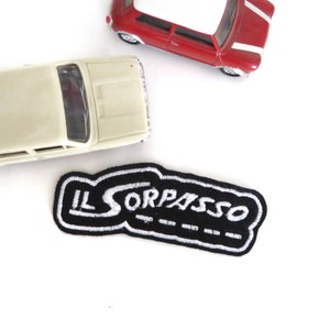 May include: A black and white embroidered patch with the text "IL SORPASSO" in a white cursive font. The patch is surrounded by a black border.
