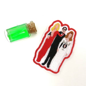 Pode incluir: Etiqueta bordada com três figuras, um homem de terno preto e duas mulheres, uma de vestido vermelho e a outra de vestido branco. A etiqueta é cercada por uma borda branca.