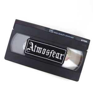 Puede incluir: Cinta VHS negra con un parche bordado blanco que dice "Atmosfear". La cinta está etiquetada como "HS High Quality Standard" y "TDK".