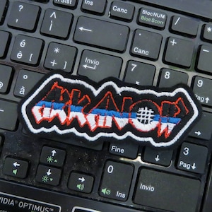 Könnte beinhalten: Ein schwarzer und weißer gestickter Patch mit dem Wort "KILLER" in roten und blauen Buchstaben. Der Patch hat einen weißen Streifen mit einem schwarzen Hashtag-Symbol.