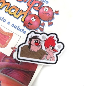 May include: An embroidered patch featuring two cartoon characters. The character on the left is a large, round man with a red nose and a brown shirt. The character on the right is a thin man with red hair and a white shirt. The patch is surrounded by a white border and has a cloud-like design behind the characters.