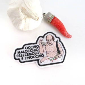 May include: Embroidered patch featuring a cartoon man with a white background. The man is holding a bowl and a knife. The text on the patch reads "OCCHIO MALOCCHIO PREZZEMOLO E FINOCCHIO".