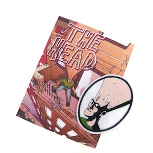 Pode incluir: Um remendo bordado branco e preto de um personagem de desenho animado com uma cabeça grande e óculos. O personagem está vestindo uma camisa verde. O remendo está em cima de uma capa de quadrinhos com o título "The Head" em letras vermelhas.