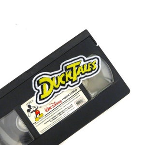 Puede incluir: Una cinta VHS negra con una pegatina amarilla y blanca con el logotipo de "DuckTales" en la parte delantera. La cinta está etiquetada como "Walt Disney Home Video" y tiene un personaje de Mickey Mouse en el lado izquierdo.