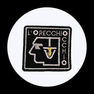 Puede incluir: Parche bordado negro con el texto blanco "L'ORECCHIO OCCHIO". El diseño incluye un perfil estilizado y el número "3" en amarillo. El parche es cuadrado con un borde blanco.