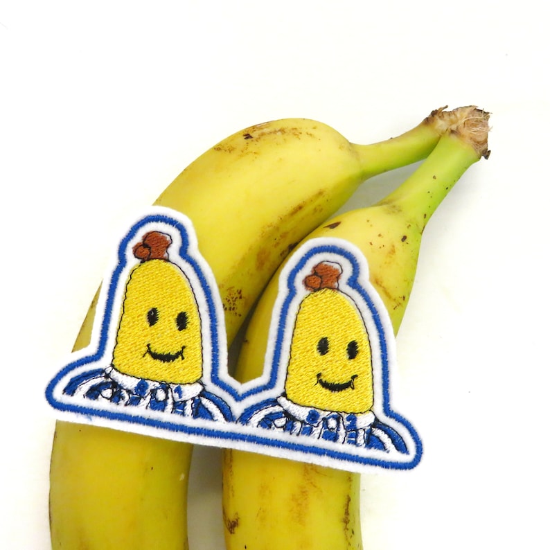 May include: Two embroidered iron-on patches of smiling yellow bananas wearing blue and white striped shirts. The patches are on top of two yellow bananas.
