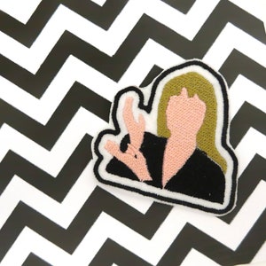Op de afbeelding: Een geborduurd strijk-patch met een cartoonfiguur met blond haar, die een zwarte jurk draagt en een hand opsteekt in een groet. De huid van het personage is roze en de achtergrond is wit.