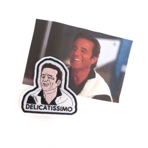 Può includere: Toppa ricamata bianca con un'immagine di un uomo cartone animato con un'espressione sorpresa. Il testo "Delicatissimo" è sotto l'immagine.