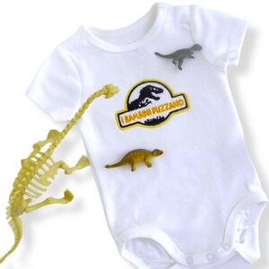 May include: White baby onesie with a yellow and orange dinosaur patch that says "I Bambini Puzzano". There are three small dinosaur toys on the onesie.