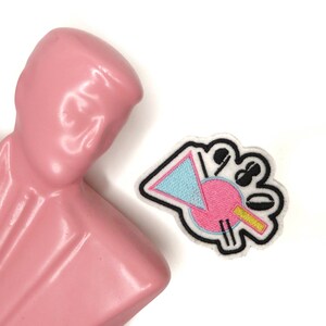 Puede incluir: Un parche bordado blanco con un diseño geométrico rosa, azul y amarillo. El parche tiene un contorno negro y está sobre un fondo blanco.