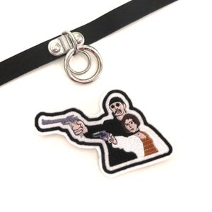 Könnte beinhalten: Ein schwarzes Lederhalsband mit einem silbernen Doppelringverschluss und einem gestickten Aufnäher mit zwei Figuren, die Waffen halten. Der Aufnäher ist weiß mit schwarzen Umrissen und zeigt einen Mann und eine Frau.