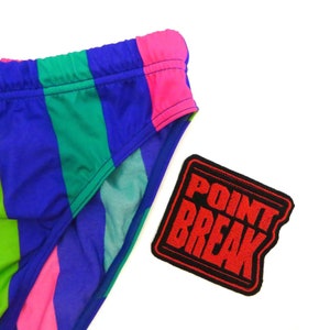 Pode incluir: Um fato de banho listrado colorido com um remendo preto que diz "POINT BREAK" em letras vermelhas.
