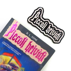 May include: A book titled "Piccoli Brividi" by R. L. Stine with a pink foil title and a black and white embroidered patch with the same title.