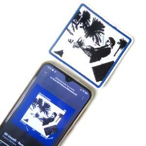 Puede incluir: Parche cuadrado blanco con un gráfico en blanco y negro de una persona y palmeras, enmarcado por un borde azul. Un smartphone muestra la portada de un álbum con el mismo gráfico y el texto "La Voce Del Padrone (Remastered)".