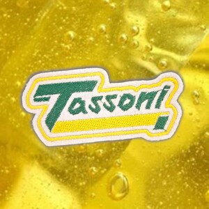 Può includere: Toppa ricamata con la scritta "Tassoni" in verde e giallo, bordata di bianco. Lo sfondo è un liquido giallo e frizzante. Adatta per abbigliamento o accessori.