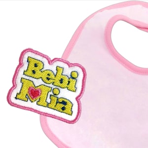 May include: Pink baby bib with a yellow and white embroidered patch that says "Bebi Mia" with a red heart.