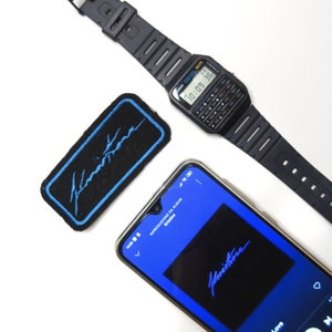 May include: A black digital watch with a calculator function, a black patch with blue stitching and the word "Kristine" in blue, and a blue smartphone screen displaying the album art for "Kristine" with the word "Kristine" in white.