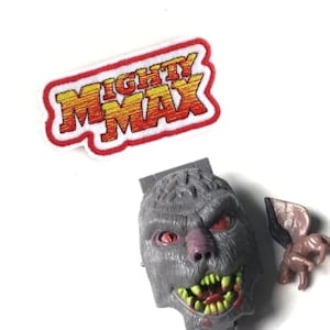 May include: A Mighty Max toy set featuring a gray monster head with red eyes and sharp teeth, a small gray and brown creature with wings, and a white patch with the words "Mighty Max" in red and yellow letters.