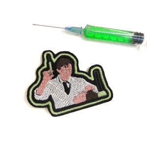 May include: An embroidered iron-on patch featuring a cartoon doctor holding a syringe and a patient in a dentist's chair. The patch has a green outline and is on a white background.