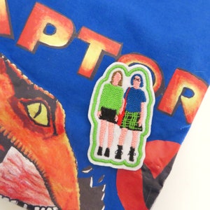 Può includere: Una toppa ricamata da stirare con due figure di donne dei cartoni animati in piedi una accanto all'altra. La donna a sinistra indossa una camicia verde, una gonna nera e scarpe nere. La donna a destra indossa una camicia blu, una gonna a quadri e scarpe nere. La toppa è su uno sfondo blu con la parola "RAPTOR" stampata in giallo e arancione.