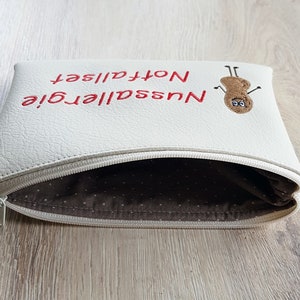 May include: Cream-colored zippered pouch with red embroidered text reading "Nussallergie Notfallset" and a peanut character. The interior lining is a brown fabric with white polka dots. The zipper is silver.