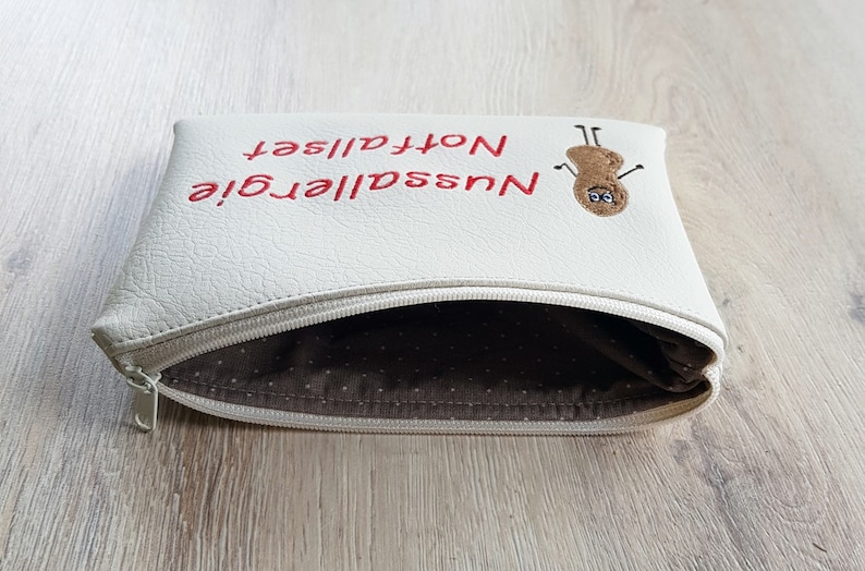 May include: Cream-colored zippered pouch with red embroidered text reading "Nussallergie Notfallset" and a peanut character. The interior lining is a brown fabric with white polka dots. The zipper is silver.
