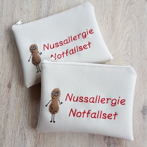 May include: Two white zippered pouches with the text "Nussallergie Notfallset" in red. Each pouch features an embroidered peanut character. The pouches are made of a textured material.