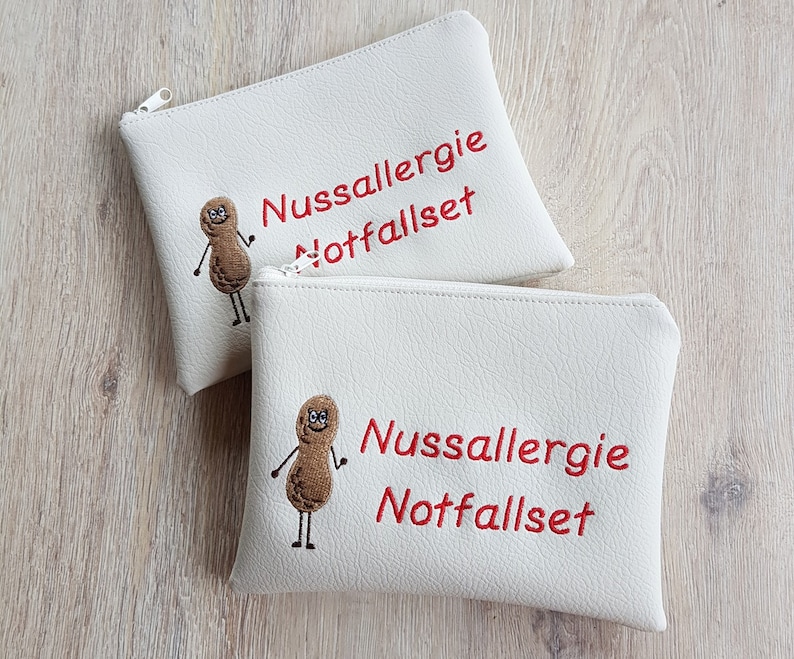May include: Two white zippered pouches with the text "Nussallergie Notfallset" in red. Each pouch features an embroidered peanut character. The pouches are made of a textured material.