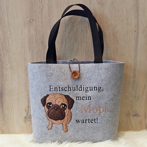 Könnte beinhalten: Graue Filztasche mit schwarzen Henkeln und einem braunen Knopf. Die Tasche hat ein gesticktes Bild eines Mops und den Text "Entschuldigung, mein Mops wartet!"