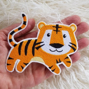Könnte beinhalten: Orangefarbener und schwarzer gestickter Bügelaufkleber mit einem Cartoon-Tiger mit weißem Gesicht und schwarzen Streifen.